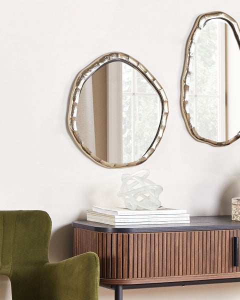 Zwei unregelmäßige Spiegel mit goldenen Rahmen hängen an der Wand über einem Sideboard mit Dekorationsgegenständen und Büchern.