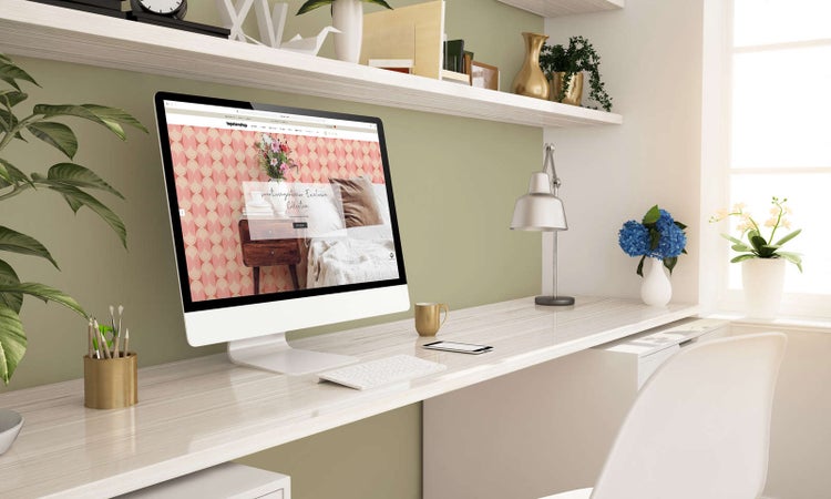 Heller und organisierter Arbeitsbereich mit einem weißen Schreibtisch in Holzoptik, einem Computerbildschirm, einer silbernen Lampe und Pflanzen.