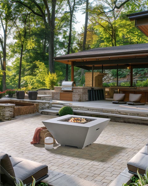 Moderne quadratische Gas-Feuerstelle auf einer gepflasterten Terrasse in einem weitläufigen Garten mit Außenküche und Wald im Hintergrund.