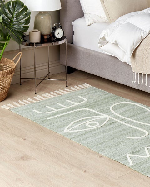 Hellgrüner Teppich mit weißem Gesichtsmotiv und Fransen auf hellem Holzboden in einem hellen Schlafzimmer mit Bett und Beistelltisch.