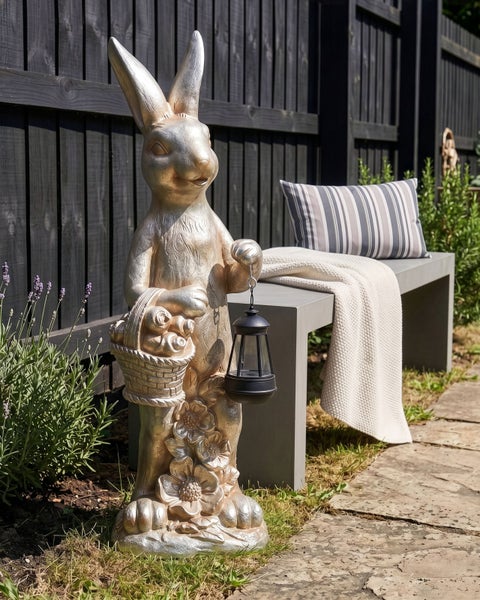 Goldfarbene Gartenfigur Hase mit Korb und schwarzer Laterne neben einer modernen Gartenbank vor dunklem Holzzaun.