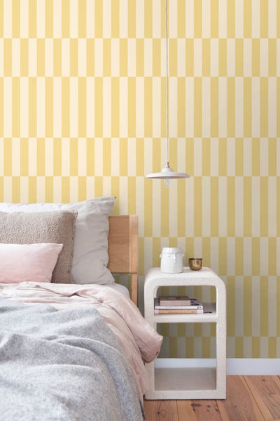 Schlafzimmer mit geometrisch gemusterter Tapete, einem Holzbett, einem weißen, abgerundeten Nachttisch und einer Hängeleuchte.