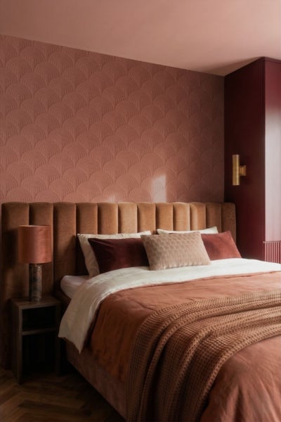 Schlafzimmer mit strukturierter Tapete, gepolstertem Kopfteil aus Samt, Bett mit mehreren Kissen, Strickdecke und Tischlampe auf dem Nachttisch.