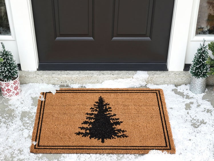 Fußmatte mit Tannenbaum-Motiv vor einer Haustür, dekoriert mit kleinen Tannenbäumen und Kunstschnee