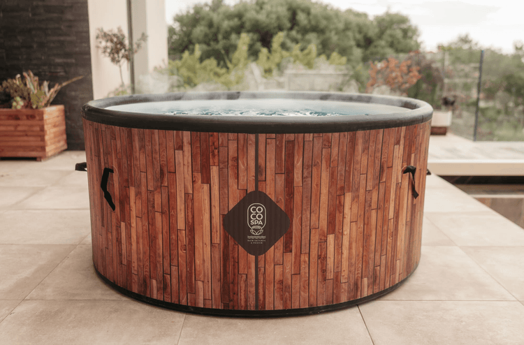 Runder aufblasbarer Whirlpool in Holzoptik mit dampfendem Wasser auf einer hellen Fliesenterrasse im Außenbereich, COCO SPA Logo.