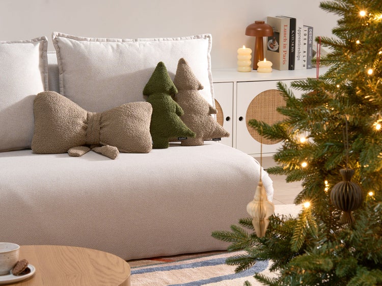 Dekoratives Wohnzimmer mit Sofa, Zierkissen in Schleifen- und Tannenbaumform sowie beleuchtetem Weihnachtsbaum