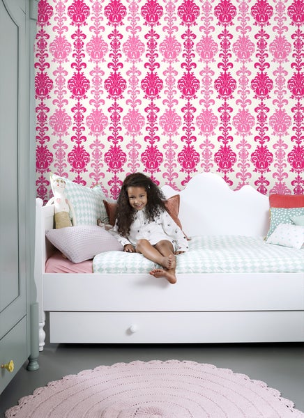 Mädchenzimmer mit weißem Bett, gemusterten Kissen und einer pinkfarbenen Mustertapete.