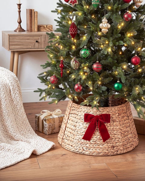 Geflochtener Korb als Weihnachtsbaumständer mit roter Samtschleife unter einem dekorierten Weihnachtsbaum im Wohnzimmer.