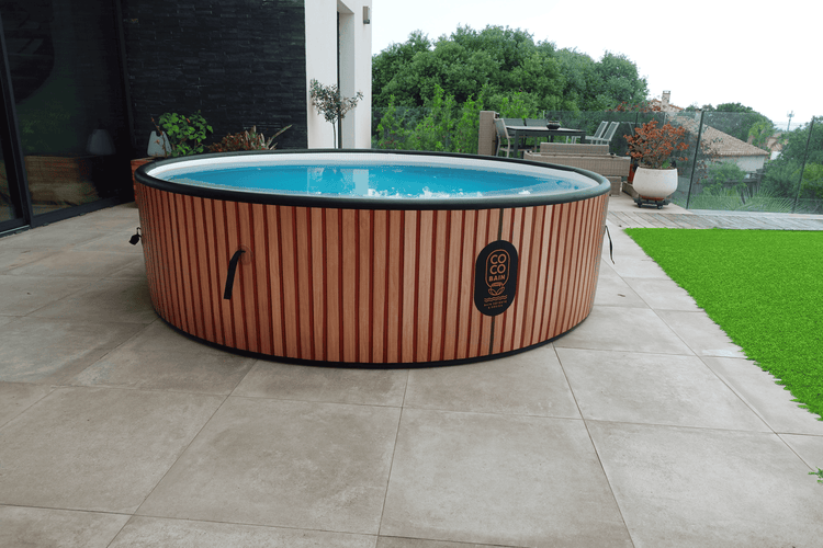 Runder aufblasbarer Whirlpool mit Verkleidung in Holzoptik auf einer hellen modern gefliesten Terrasse mit Coco Bain Logo.