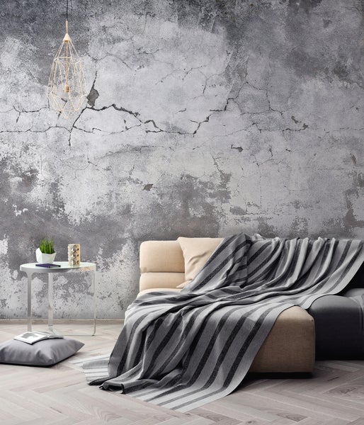 Hell gestaltetes Wohnzimmer mit Vliestapete in Betonoptik, Sofa mit gestreifter Decke, geometrischer Leuchte und Boden in Fischgrätoptik.