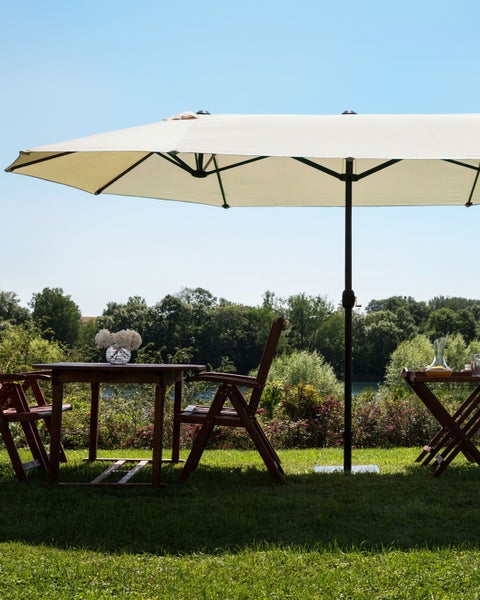 Großer rechteckiger Sonnenschirm spendet Schatten über einem hölzernen Gartentisch und Stühlen auf einer Wiese mit Blick ins Grüne.