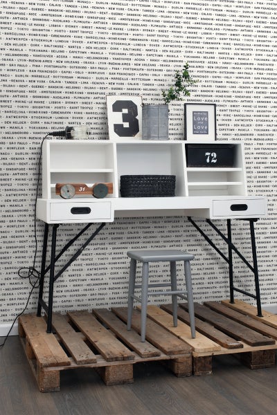 Schreibtisch mit Regalen und schwarzem Metallgestell, dazu ein grauer Hocker auf einer Holzpalette vor einer mit Städtenamen bedruckten Tapete.