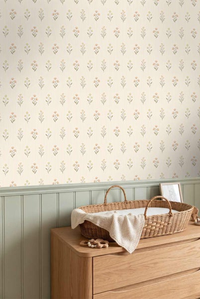 Tapete mit Blumenmuster in einem Kinderzimmer über einer grünen Wandvertäfelung und einer Holzkommode mit Wickelkorb.