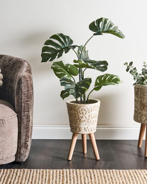 Dekorative Monstera Kunstpflanze im Topf auf Holzfüßen, neben einem Sessel auf einem dunklen Holzboden.