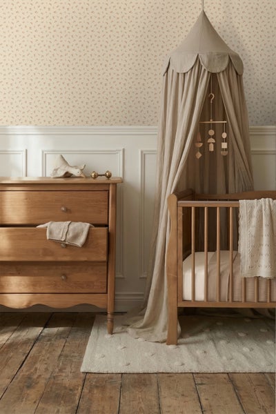 Helles Kinderzimmer mit Holzkinderbett, Baldachin, Holzkommode und floral gemusterter Tapete über weißen Wandpaneelen auf Dielenboden.