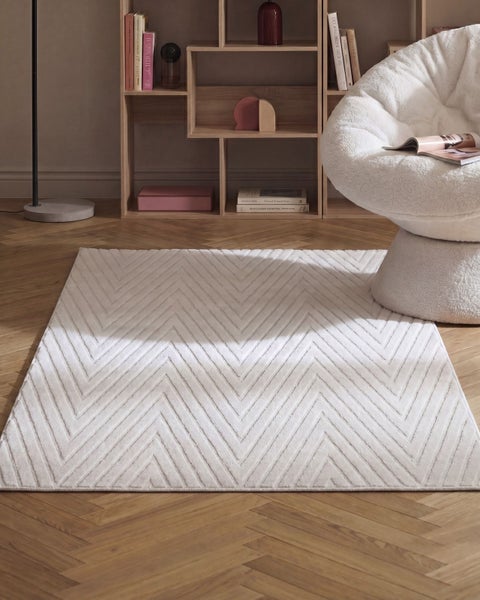 Wohnzimmer mit einem rechteckigen Teppich mit Zickzackmuster auf einem Parkettboden vor einem Regal und einem weichen Sessel.