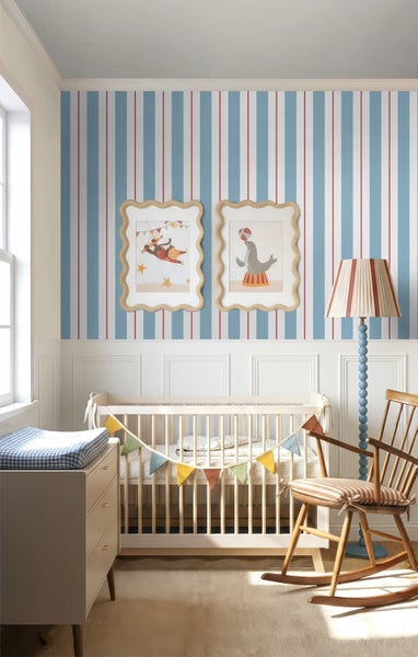 Hell eingerichtetes Kinderzimmer mit blau-weiß gestreifter Tapete, hellem Gitterbett, hölzernem Schaukelstuhl und Wickelkommode.