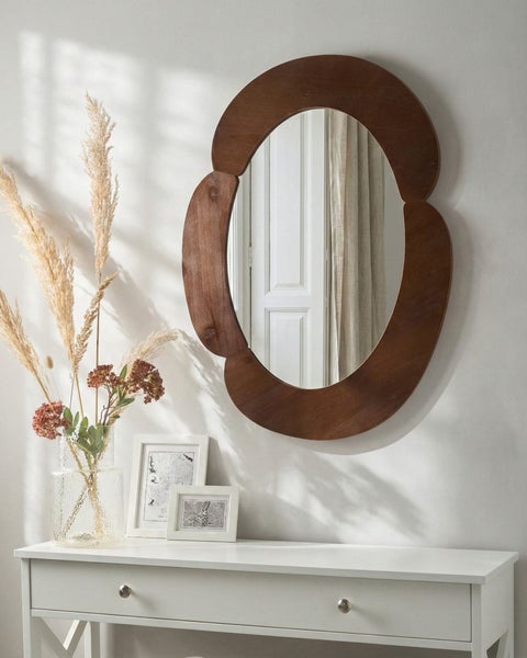 Ovaler Wandspiegel mit einem dekorativen, geschwungenen Rahmen aus dunklem Holz, montiert über einem weißen Sideboard.