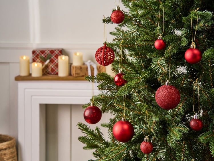 Weihnachtsbaum mit roten Kugeln in verschiedenen Designs wie Glitzer und Matt, im Hintergrund ein Kaminsims mit Kerzen und Geschenken.