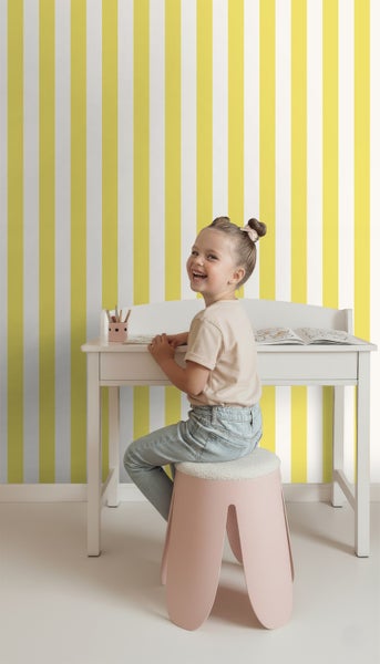 Mädchen sitzt an einem weißen Kinderschreibtisch vor einer gelb-weiß gestreiften Tapete.