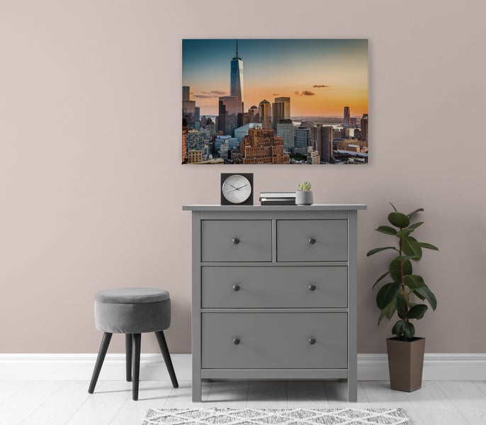 Ein Wandbild mit einer Skyline von New York City bei Sonnenuntergang hängt über einer grauen Kommode in einem modern eingerichteten Raum.