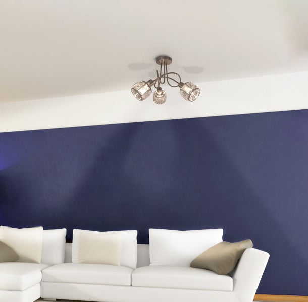 Deckenleuchte mit drei Leuchten und dekorativen Glaselementen über einem weißen Sofa vor einer blauen Wand.