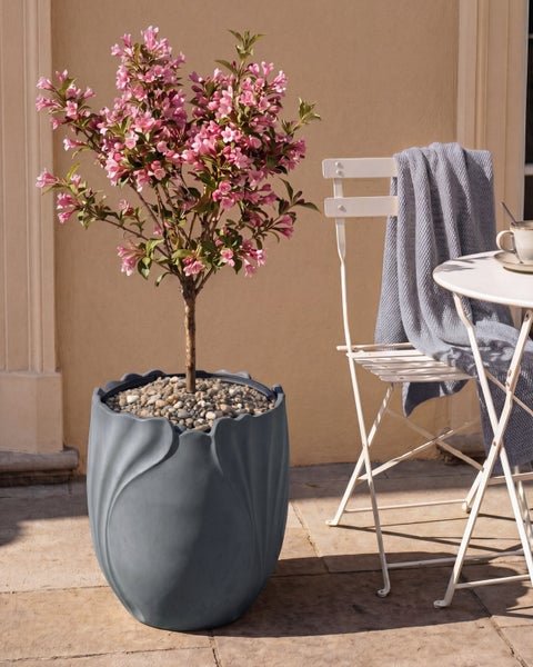 Blühende Weigela in einem grauen Pflanzkübel mit organischer Struktur neben weißem Klappstuhl und Tisch auf einer sonnigen Terrasse.