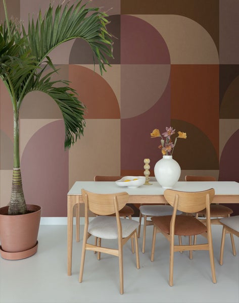 Ein Esszimmer mit Esstisch, Stühlen und einer Zimmerpflanze vor einer gemusterten Tapete.