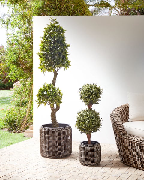 Szene mit zwei künstlichen Formgehölzen in geflochtenen Pflanzgefäßen und einem Korbsessel auf einer Terrasse.