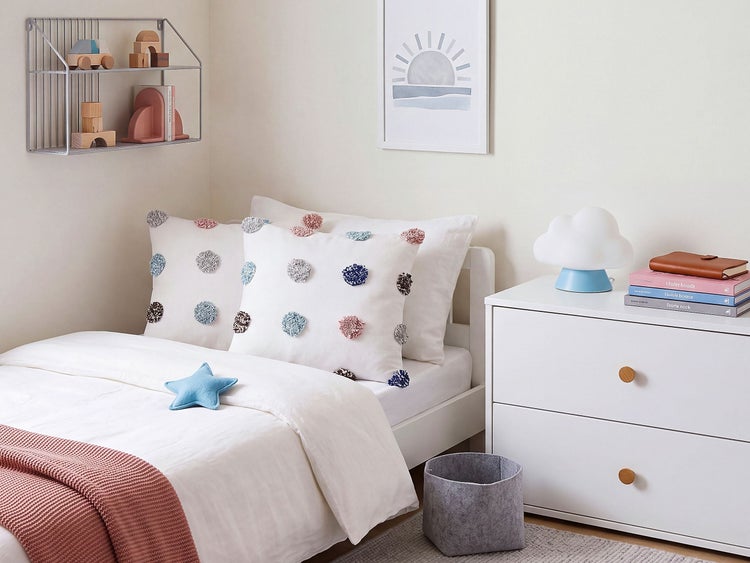 Ein helles Kinderzimmer mit weißem Bett, gepunkteten Kissen, einer Wolkenlampe auf einer weißen Kommode und Wandregal mit Holzspielzeug.