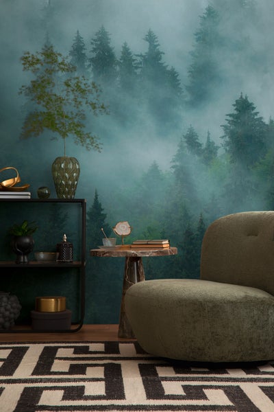 Fototapete mit nebligem Nadelwald in einem Wohnzimmer mit grünem Sessel, Marmortisch, Regal und gemustertem Teppich.