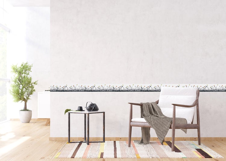 Helles Wohnzimmer mit floraler Tapetenbordüre, Holzarmsessel mit Decke, Beistelltisch und gestreiftem Teppich auf Holzboden.