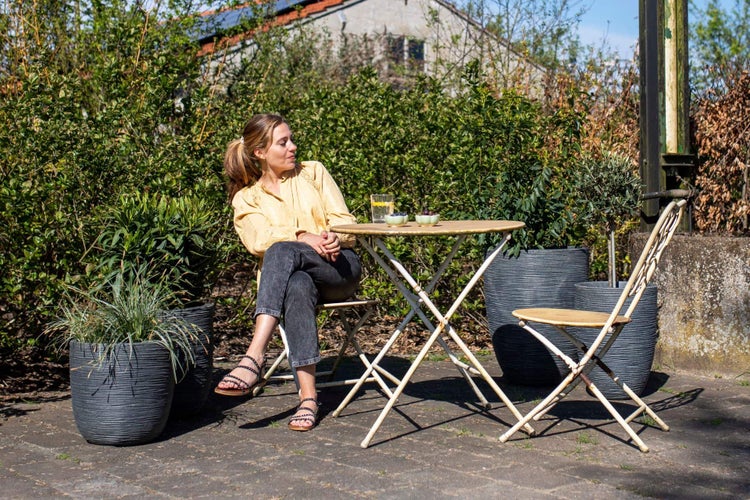 Frau sitzt an einem runden Gartentisch mit Klappstühlen auf einer Terrasse, umgeben von großen anthrazitfarbenen Pflanzkübeln.