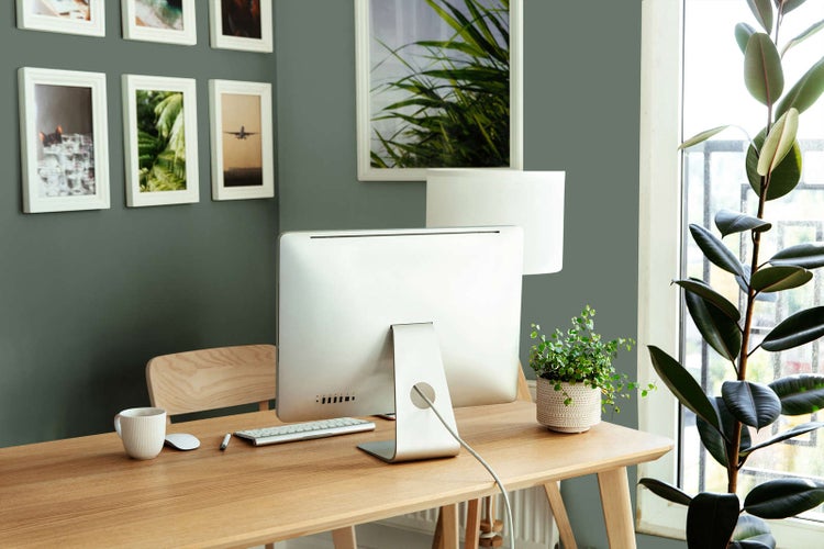 Moderner Heimarbeitsplatz mit Holzschreibtisch, Computer, Zimmerpflanzen und Bilderrahmen an einer grünen Wand.