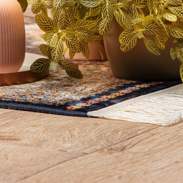 Teppich mit orientalischem Muster, dunklem Rand und weißen Fransen auf einem hellen Boden in Holzoptik neben einer Zimmerpflanze.