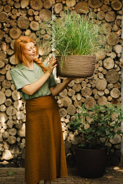 Frau mit geflochtenen Haaren hält einen runden Pflanztopf mit Gras vor einer Wand aus Holzstämmen.