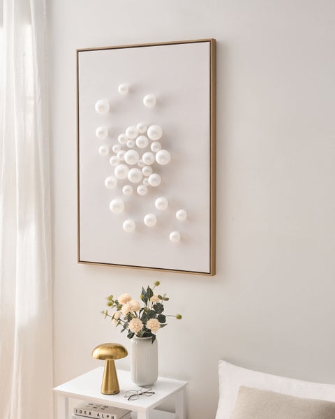 Modernes Wandbild mit dreidimensionalen weißen Kugeln in hellem Rahmen, über einem Beistelltisch mit Lampe und Vase in hellem Raum.