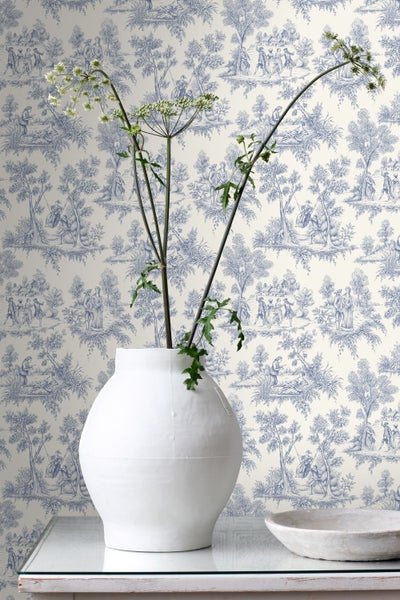 Vliestapete mit blauem Toile-de-Jouy-Muster, das ländliche Szenen zeigt, kombiniert mit einer weißen Vase und getrockneten Blumen im Vordergrund.