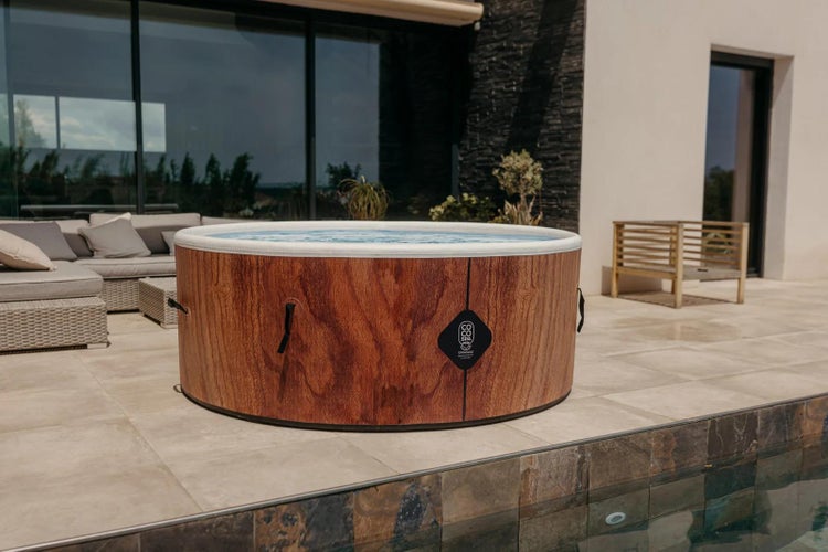 Runder Außen-Whirlpool in Holzoptik auf einer gefliesten Terrasse neben einem Pool mit grauen Loungemöbeln und Hausfassade im Hintergrund.