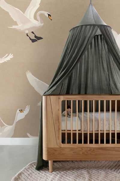 Kinderzimmer mit Holzgitterbett und grünem Baldachin vor einer Tapete mit Schwanmotiven.