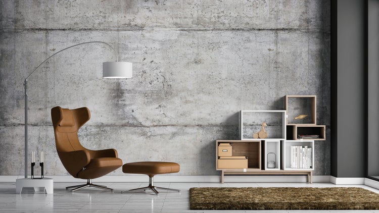 Wohnzimmer mit Betonoptik-Wand, braunem Ledersessel, passendem Hocker, moderner Stehlampe und einem modularen Regalsystem.