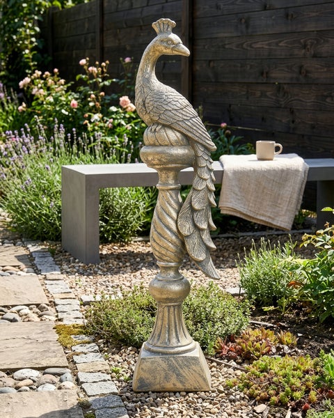 Gartenfigur Pfau auf einer dekorativen Säule in einem modern gestalteten Garten mit Steinweg, Kies, Lavendel und einer Betonbank.