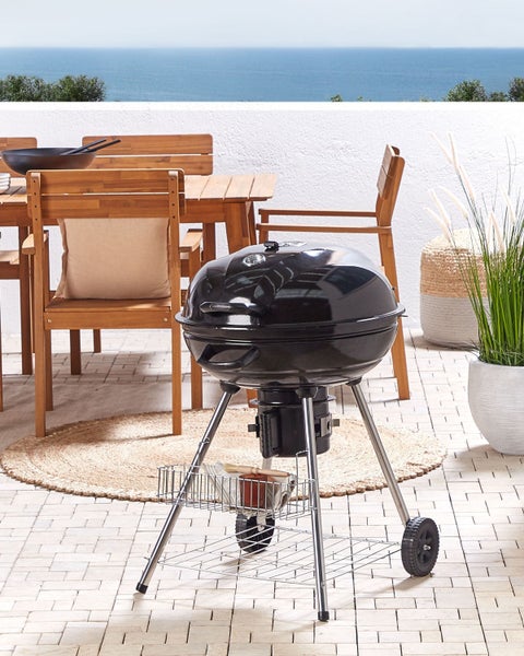 Schwarzer Holzkohle-Kugelgrill mit Thermometer und Rädern auf einer Terrasse mit Holzmöbeln und Meerblick.