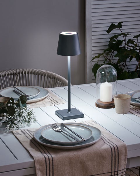 Moderne kabellose LED-Tischleuchte auf einem gedeckten Esstisch mit Tellern, Besteck und einer Kerze unter einer Glasglocke.