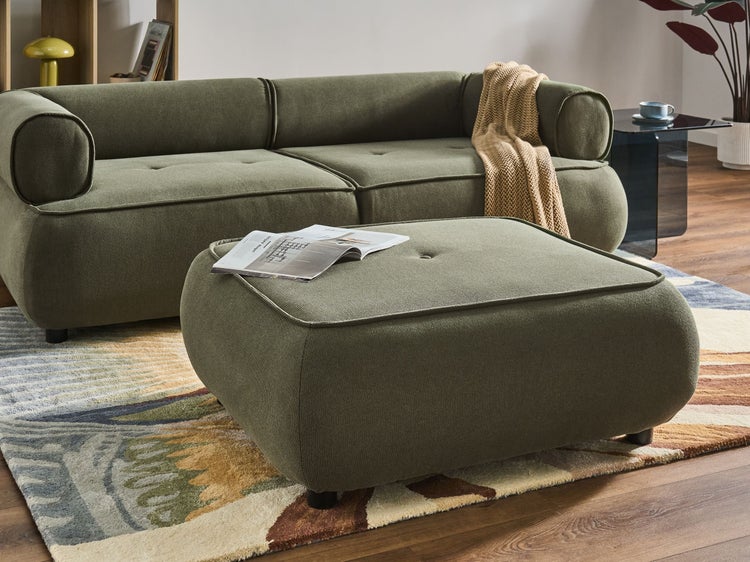 Modernes Wohnzimmer mit Polstersofa, passendem Hocker aus strukturiertem Stoff und Beistelltisch auf einem gemusterten Teppich.