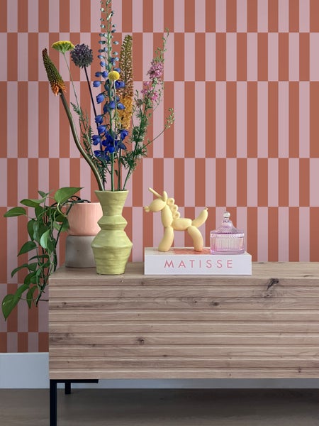 Dekoratives Stillleben mit Kommode, Blumen, Ballonfigur und Buch vor gestreifter Tapete