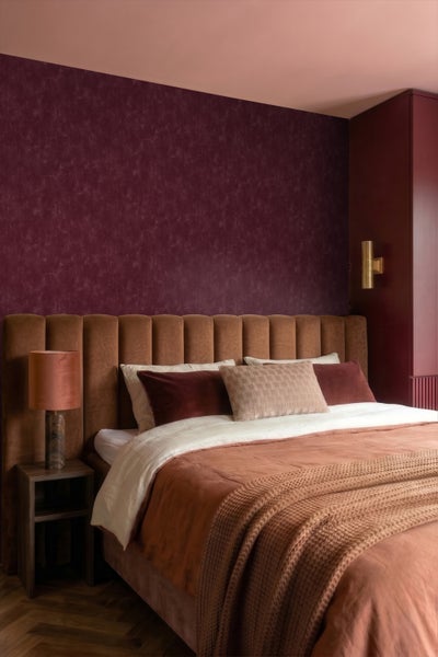 Modernes Schlafzimmer mit gepolstertem Kopfteil, strukturierter burgunderroter Tapete, verschiedenen Kissen und einer stilvollen Nachttischlampe.