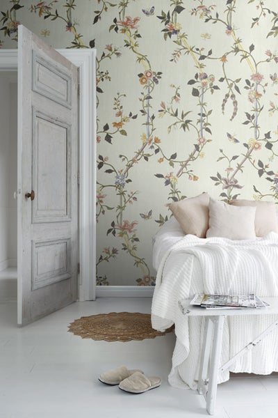 Gemütliches Schlafzimmer mit floraler Tapete, offenstehender Holztür und weißer Tagesdecke.