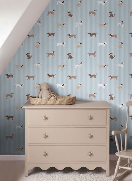 Kinderzimmer mit hellblauer Mustertapete mit Hunden, beiger Kommode mit Sternknöpfen, Weidenkorb mit Stoffhase und weißem Schaukelstuhl.