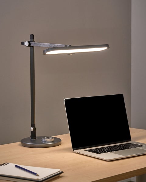 Moderne LED-Schreibtischleuchte mit verstellbarem Arm und ovalem Kopf, leuchtend auf einem Holztisch neben einem Laptop und Notizbuch.